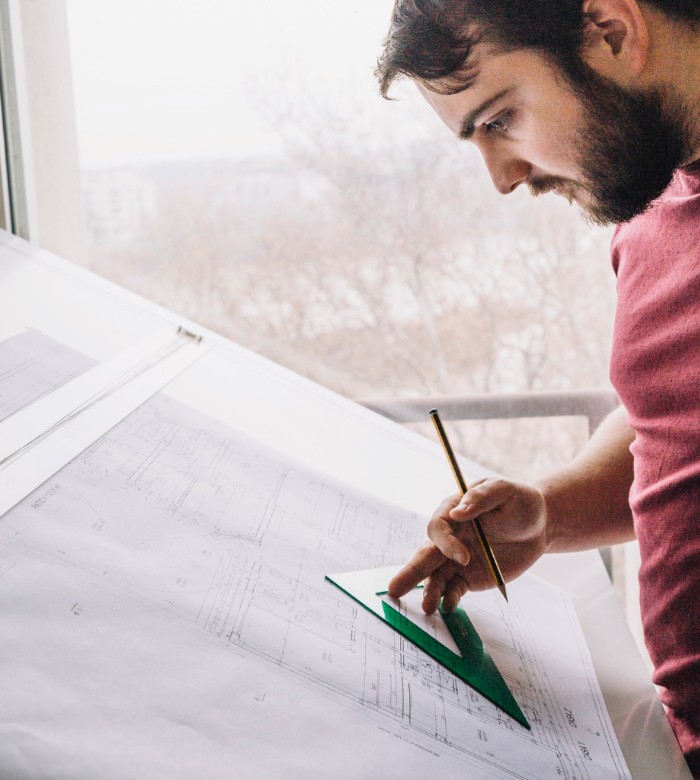 Homme étudiant un plan de bâtiment avec une équerre et un crayon.